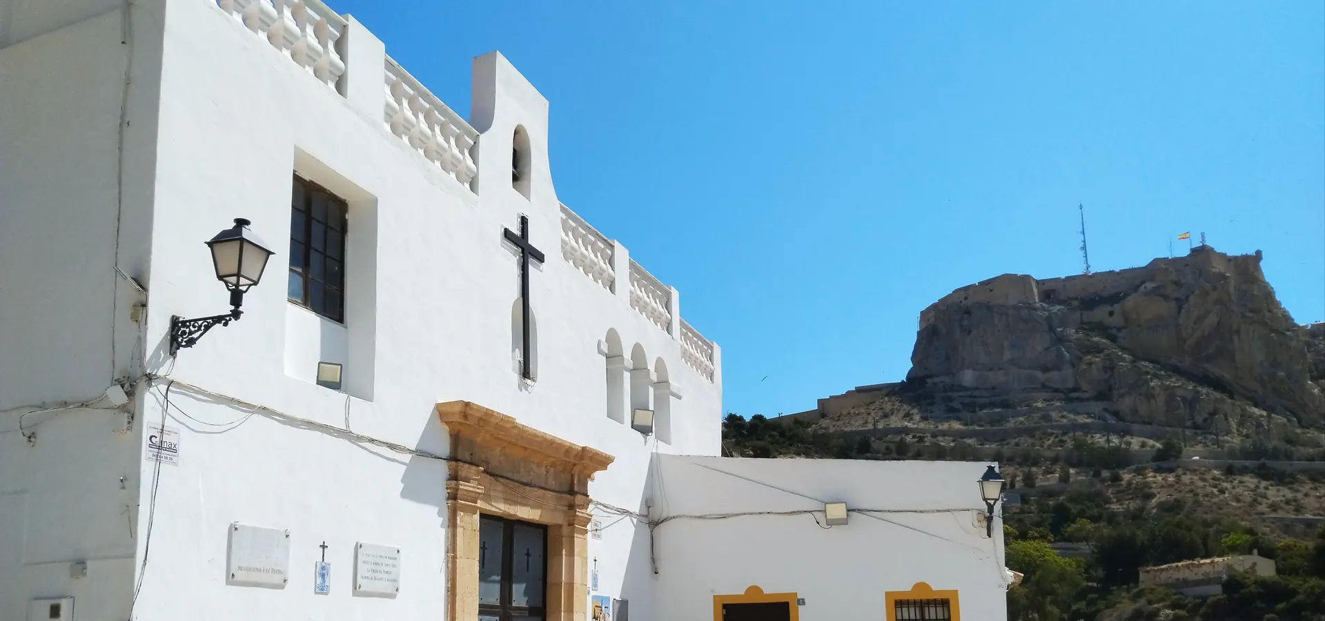 La bianca Ermita de Santa Cruz, con una croce nera sulla facciata e il basamento dipinto di giallo, e il castello sulla sua collina sullo sfondo; una fotografia generale dell'eremo in cima al quartiere