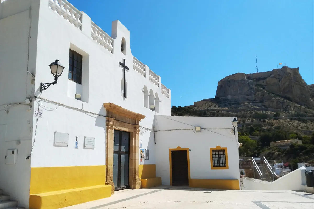 The white Ermita de Santa Cruz with a black cross on its facade and a yellow plinth, and the castle on its hill beyond