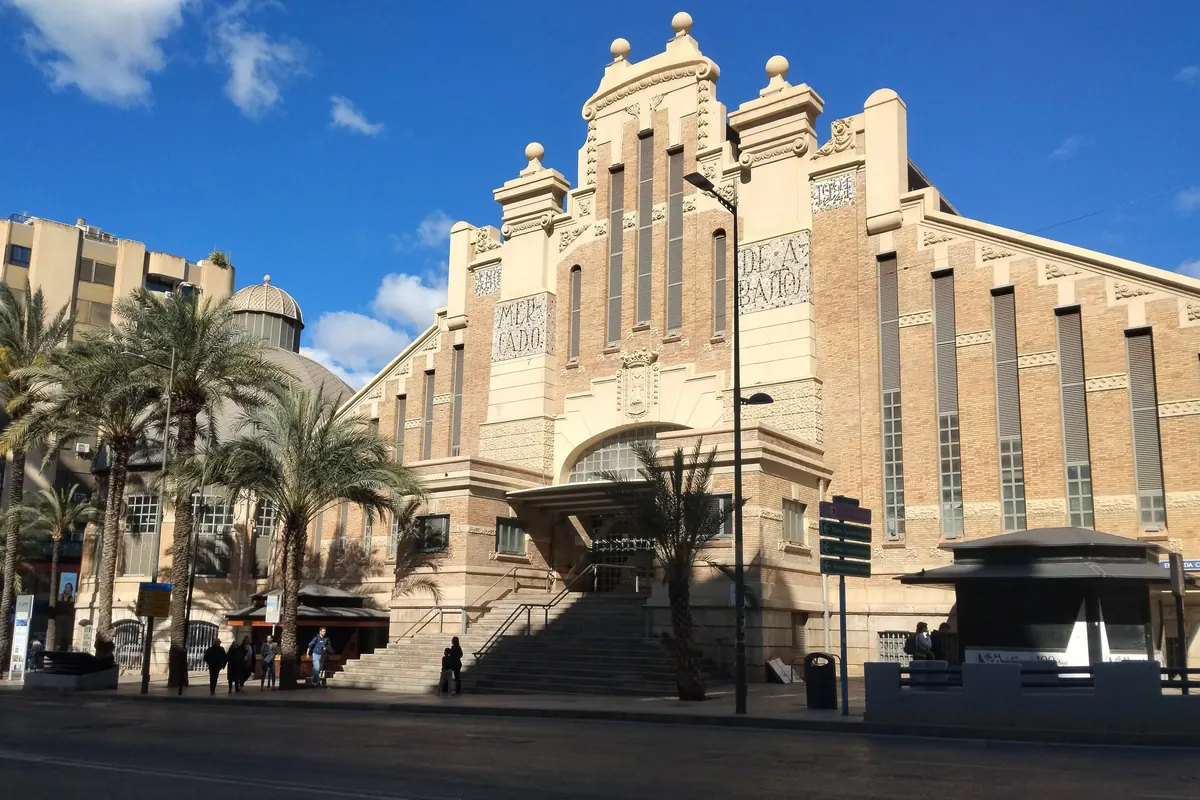 The tall cream-coloured facade of the Mercado Central de Alicante under a blue sky, with palm trees in front