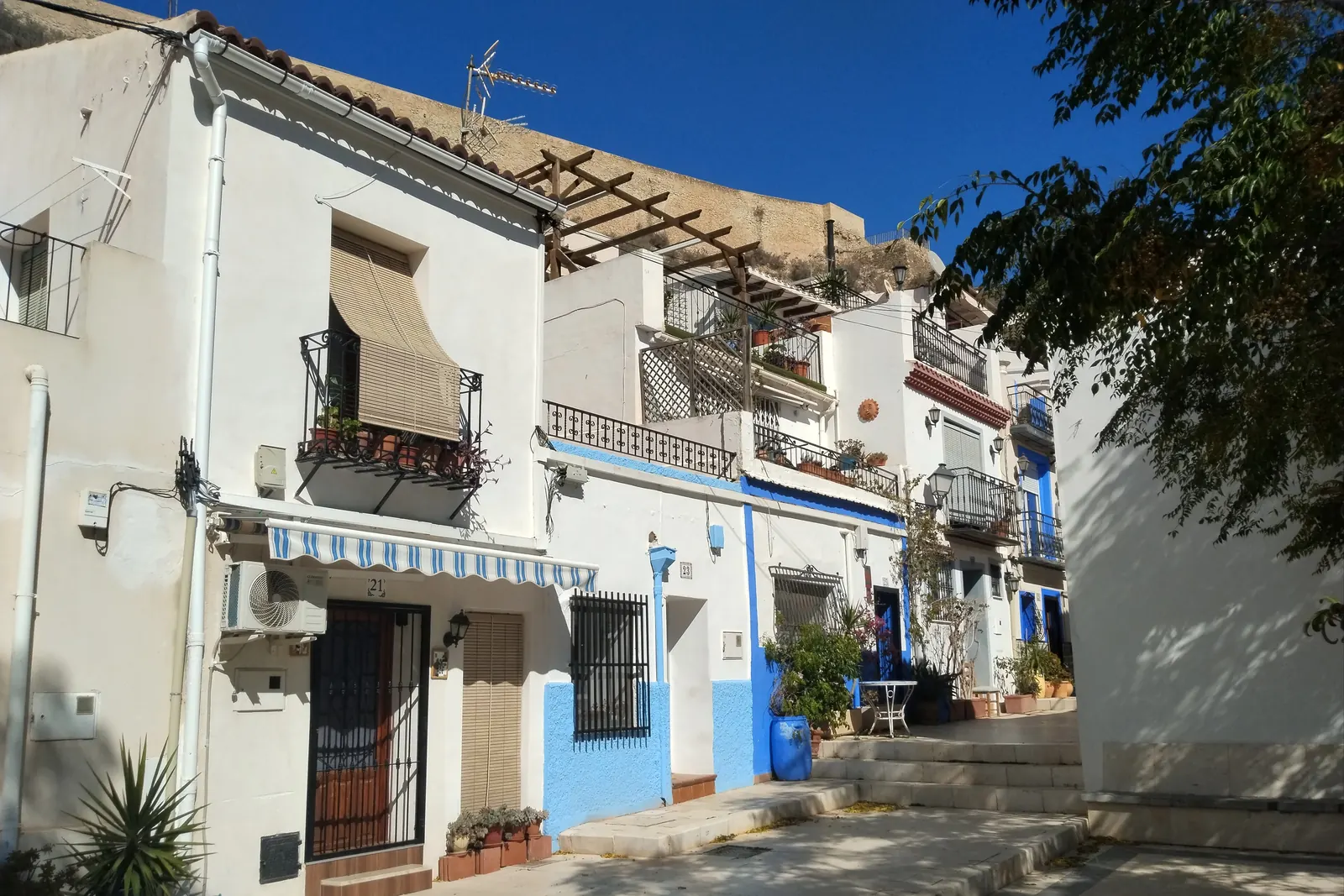 Case bianche con zoccolatura blu e persiane verdi in Calle San Antonio a Santa Cruz, con sopra la rocca della collina del castello; una fotografia generica del quartiere